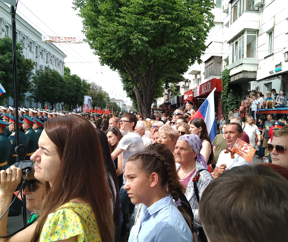 Парад начался, люди стоят в четыре ряда. На заднем фоне на шеи у мужчины сидит маленький мальчик в военном берете и с российским флагом